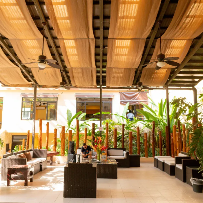 Patio techado con sof&aacute;s, plantas tropicales y ventiladores de techo. Dos personas conversan.