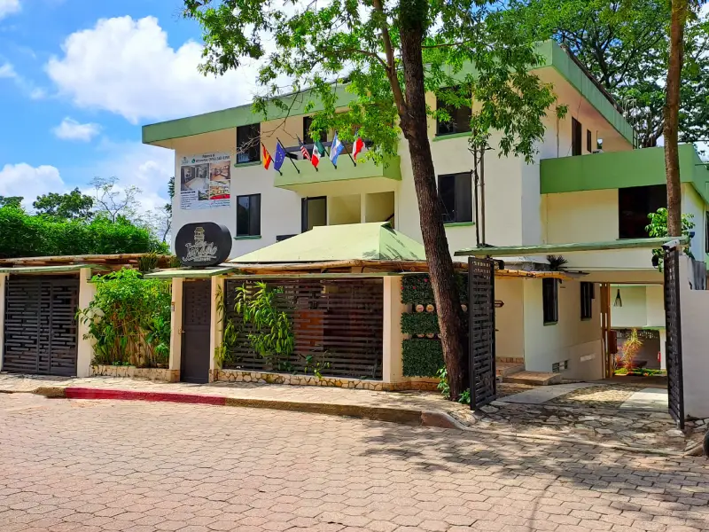 Edificio blanco y verde con banderas en el frente. &Aacute;rbol y calle adoquinada enfrente.