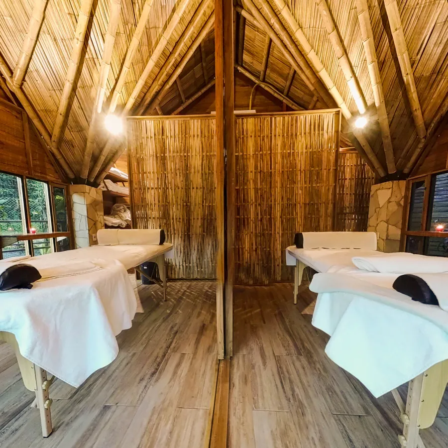 Cabaña con techo de madera y dos camas de masaje separadas por un biombo de cañas.