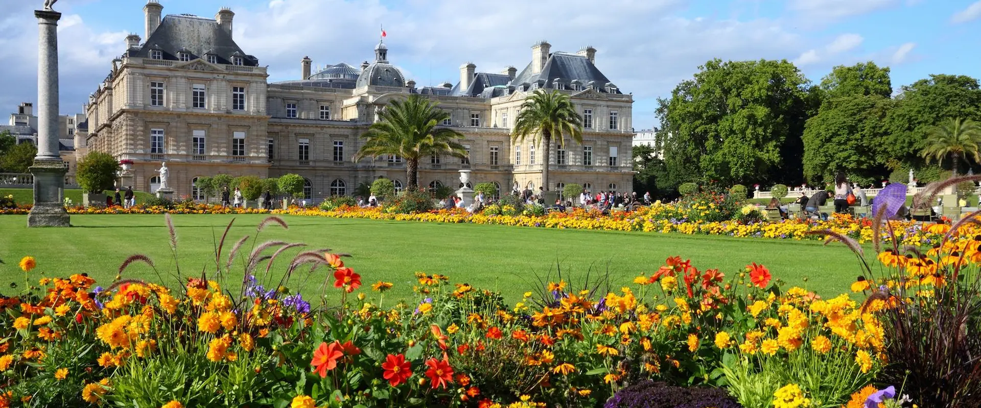 Jardin fleuri avec pelouse et grand bâtiment historique sous ciel bleu.