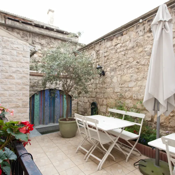 Petite cour en pierre avec table blanche, chaises pliantes et grand parasol fermé.