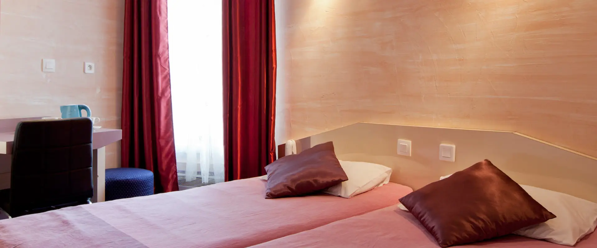 Deux lits avec couvre-lits roses, coussins marron et rideaux rouges, bureau avec chaise.