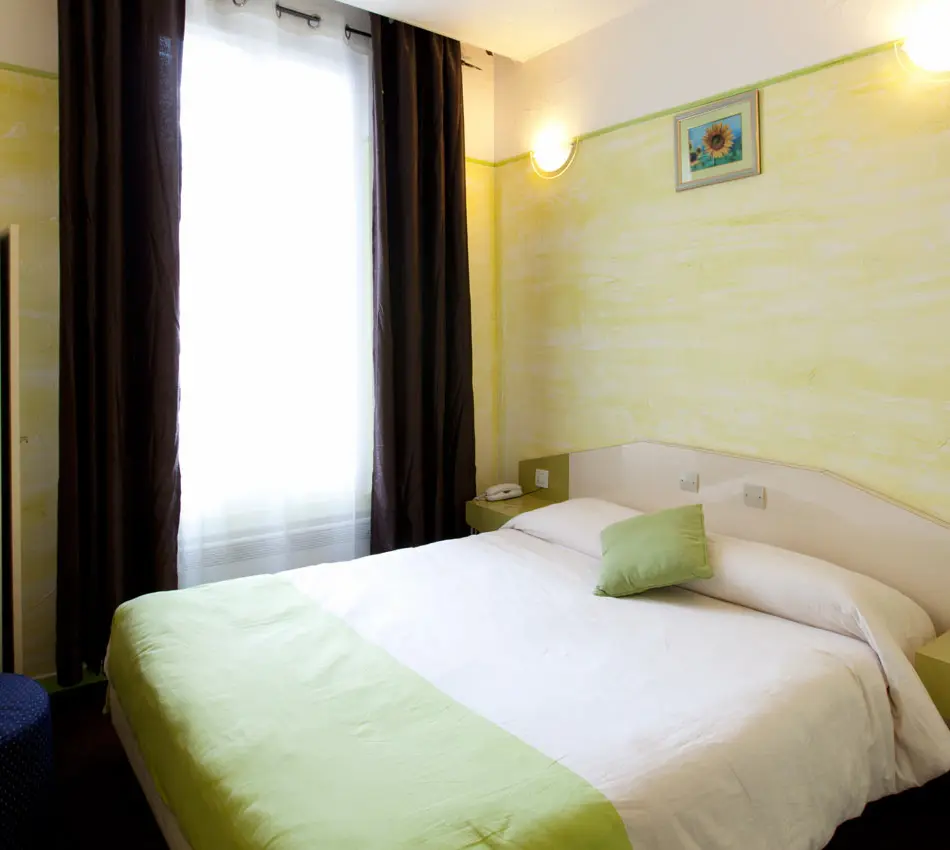 Chambre lumineuse avec un lit double, décor jaune et vert, fenêtre avec rideaux sombres.