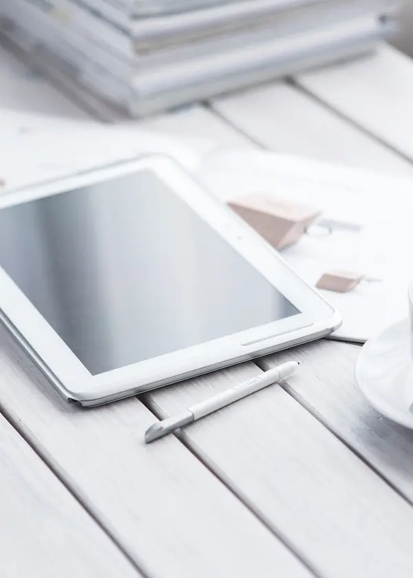 Tablette blanche avec un stylet sur une table en bois clair, près d'une tasse et de documents.