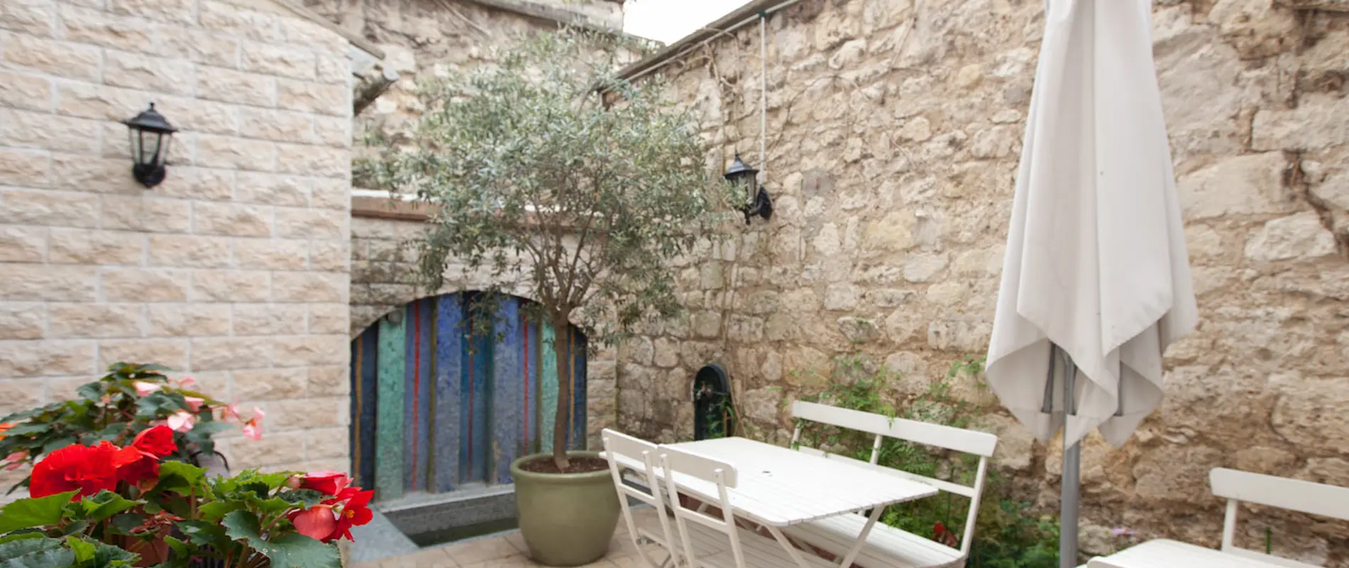 Cour intérieure en pierre avec table, chaises blanches, fleurs rouges et parasol fermé.