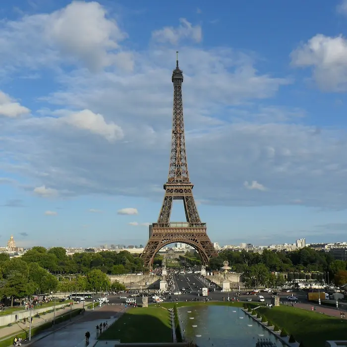 Tour Eiffel devant un ciel bleu avec des nuages épars, arbres et fontaine au premier plan.