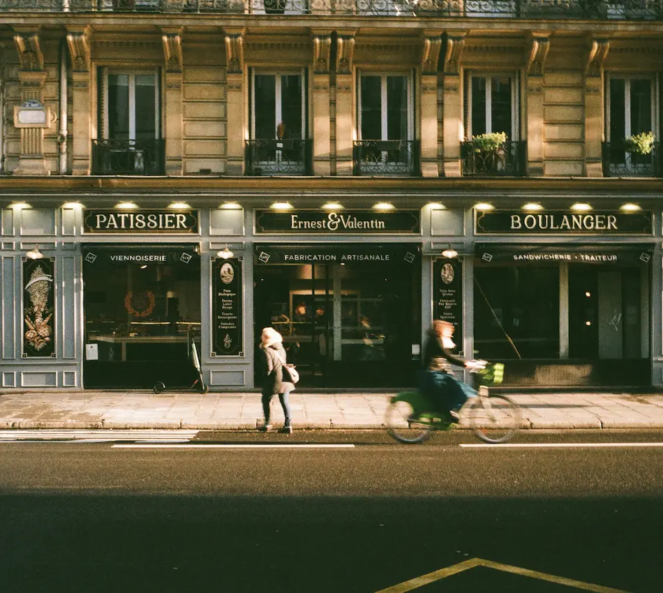 Façade d'une boulangerie-pâtisserie en ville, avec une cycliste floue et une piétonne devant.