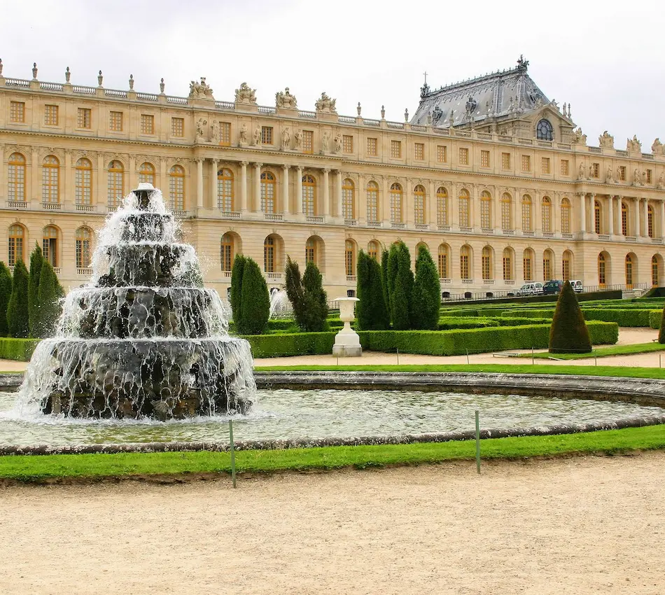 Une fontaine devant un grand palais avec des jardins soignés en arrière-plan.