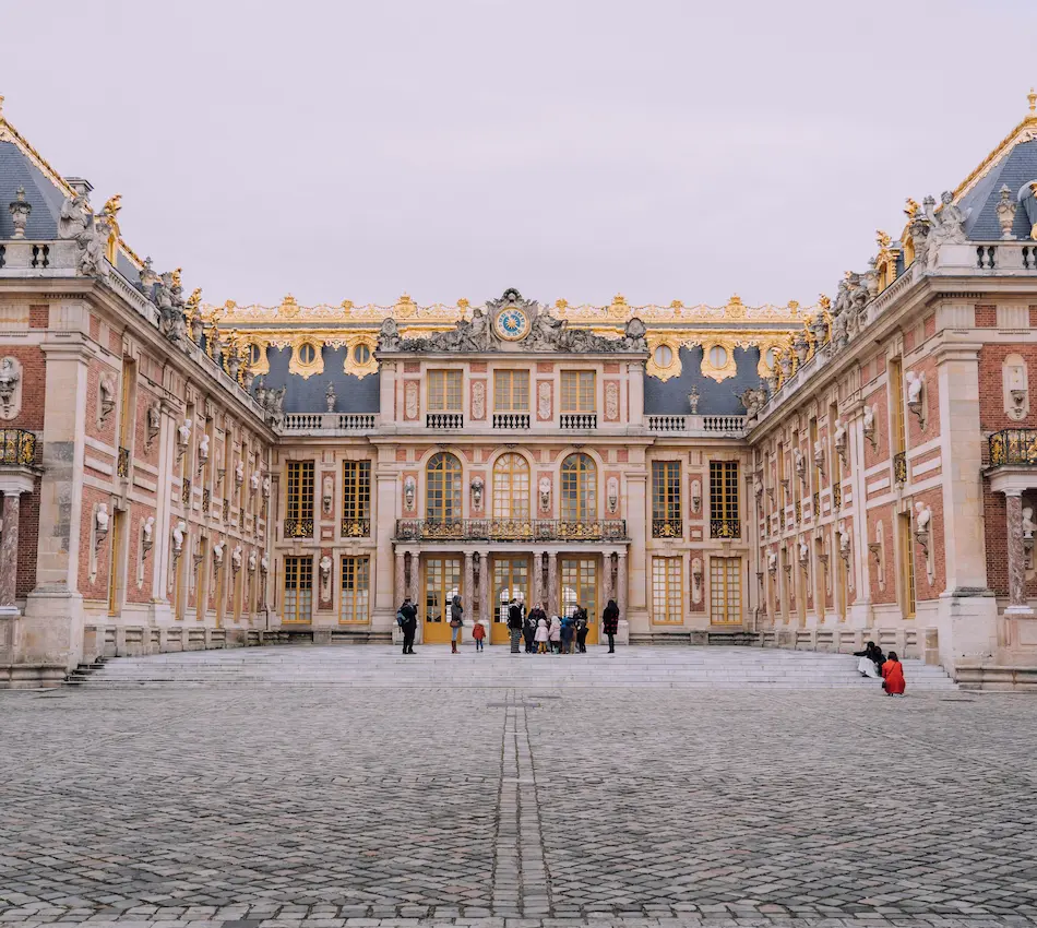 Façade du château de Versailles avec des visiteurs et une cour pavée.