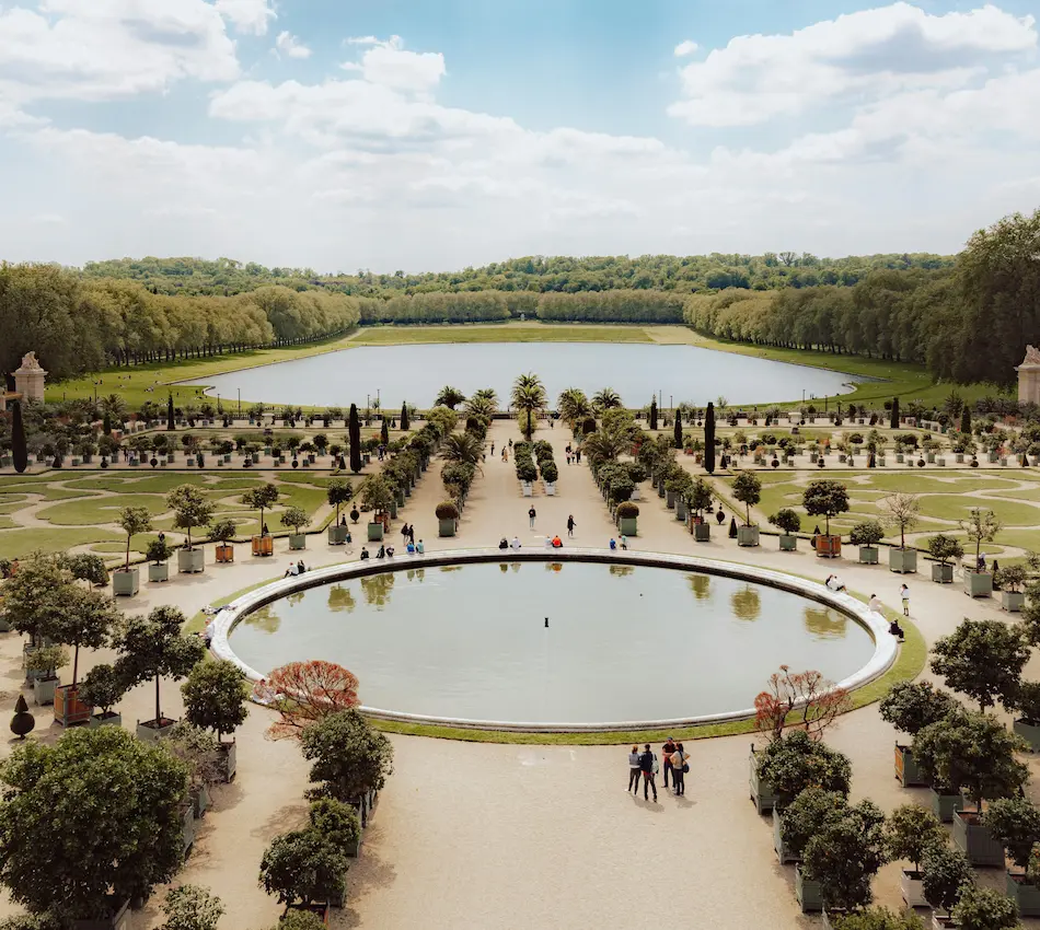 Vue d'un jardin avec fontaine et allées bordées d'arbres, ciel partiellement nuageux.