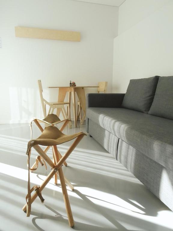 Sala minimalista com sofá cinza, banco dobrável de madeira e pequena mesa com duas cadeiras. Luz natural.