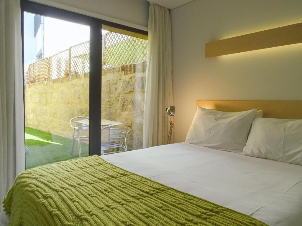 Quarto de hotel com cama de casal, cobertor verde e porta de vidro para área externa com mesa e cadeiras.