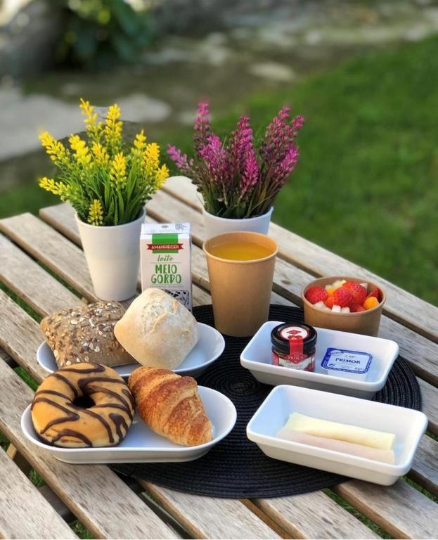 Café da manhã ao ar livre com pães, croissant, donut, frutas, suco e flores decorativas em mesa de madeira.
