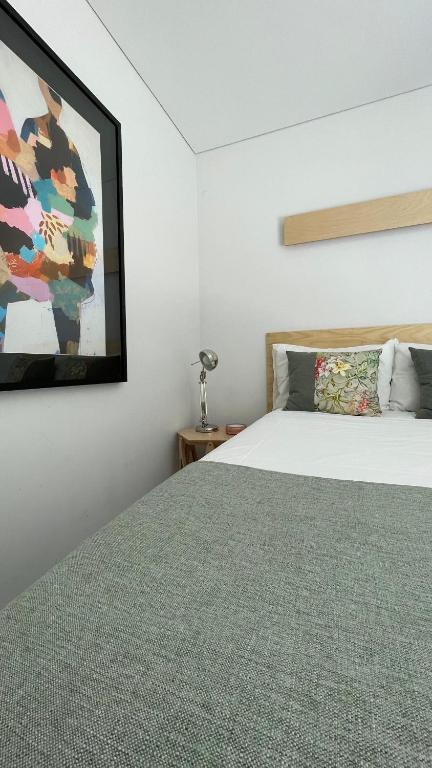 Quarto minimalista com cama de casal, colcha cinza, almofadas decorativas, mesa lateral e quadro colorido na parede.