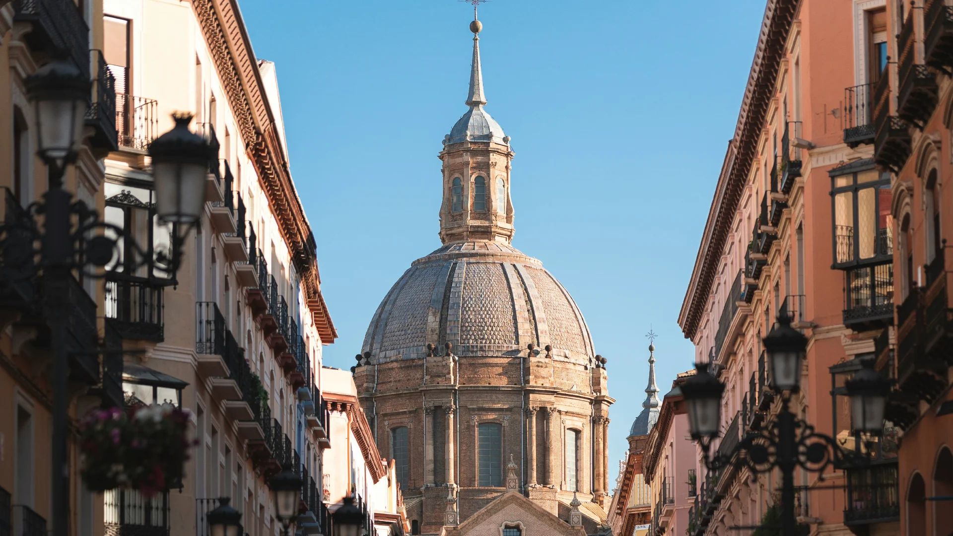 C&uacute;pula de edificio hist&oacute;rico entre edificios y faroles, con cielo claro de fondo.