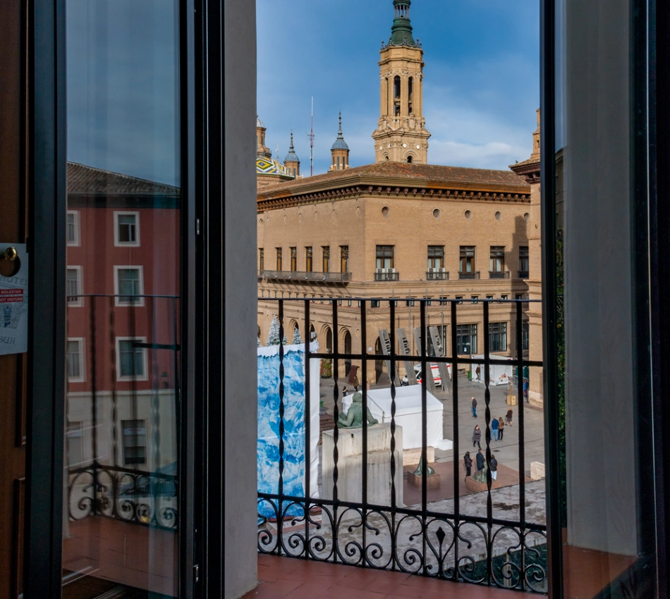 Vista desde un balc&oacute;n hacia una plaza con un edificio hist&oacute;rico y una torre al fondo.