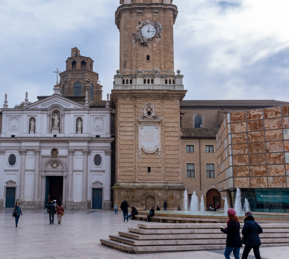 Plaza con iglesia, torre de reloj y fuente, varias personas caminando en el &aacute;rea.