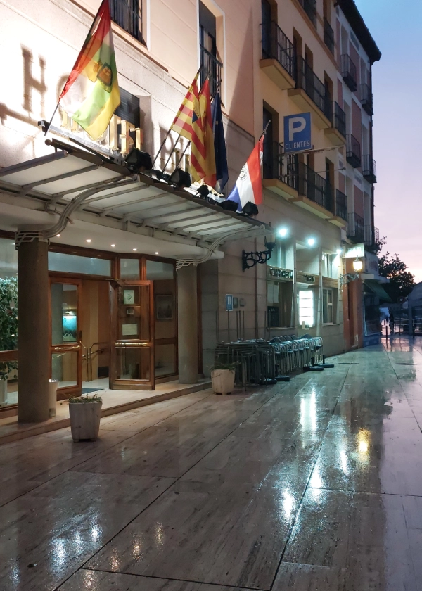 Edificio de hotel con banderas y se&ntilde;al de estacionamiento, suelo mojado reflejando luces.