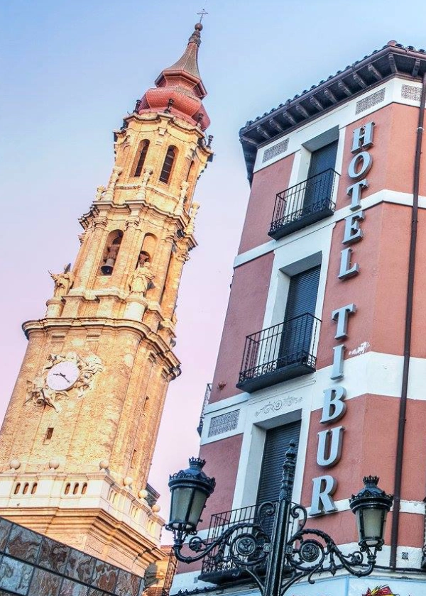Torre de iglesia junto al edificio del Hotel Tibur en una calle al atardecer.
