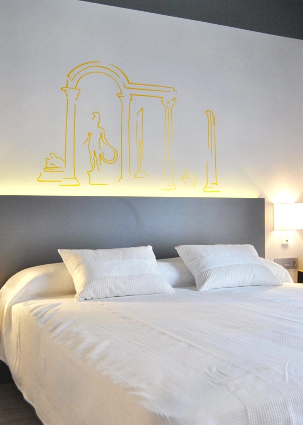 Cama blanca con luces LED y dibujo minimalista de columnas en la pared.