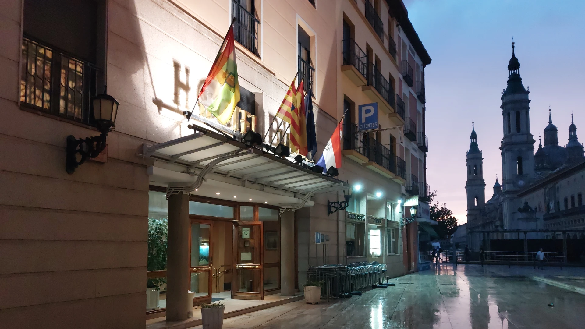 Edificio con varias banderas en la entrada, junto a una catedral al atardecer.
