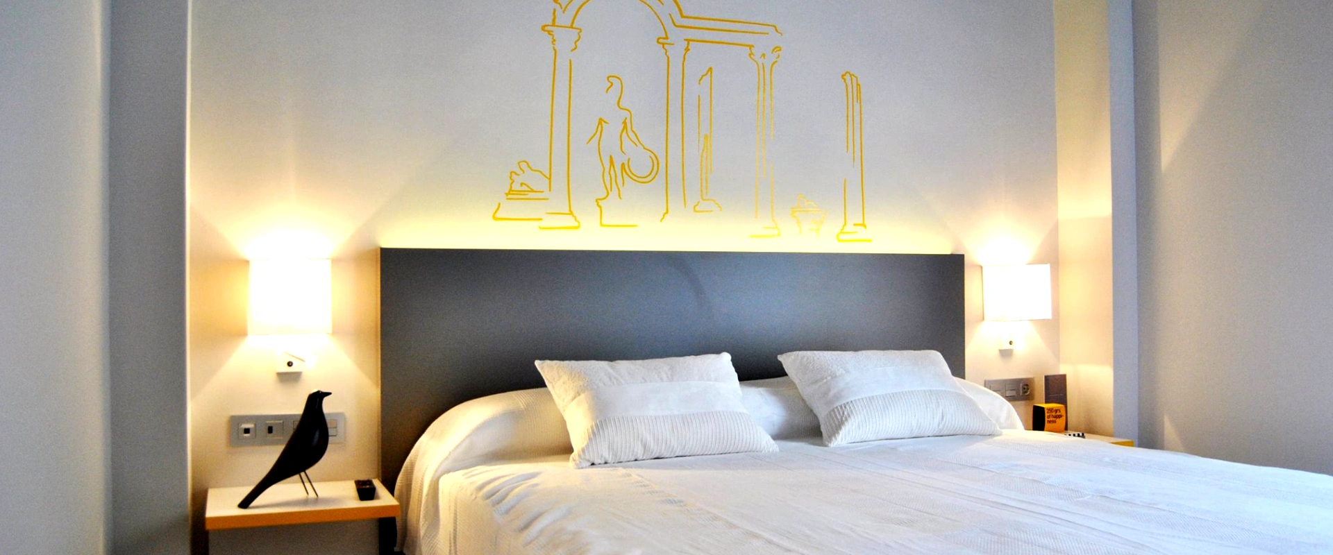 Habitaci&oacute;n moderna con cama grande, luces laterales y dibujo de columnas sobre el cabecero.