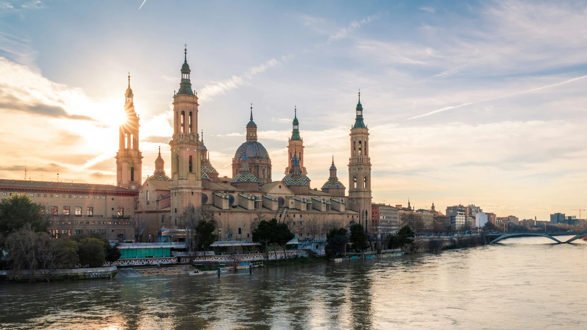 Bas&iacute;lica junto al r&iacute;o bajo un cielo al atardecer, con el sol brillando entre las torres.