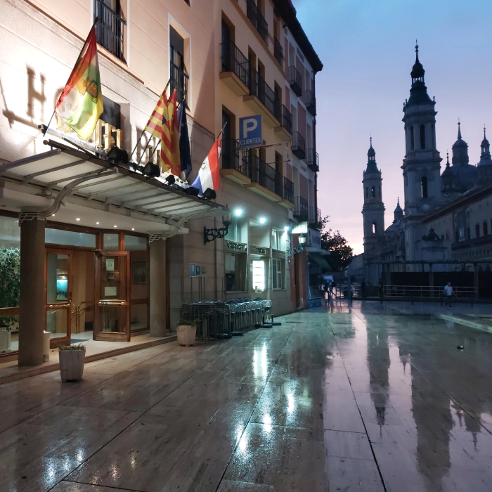 Edificio al atardecer, con banderas y suelo mojado; iglesia al fondo.