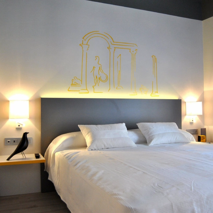 Cama doble con ropa blanca y mural amarillo de columnas cl&aacute;sicas sobre el cabecero gris.