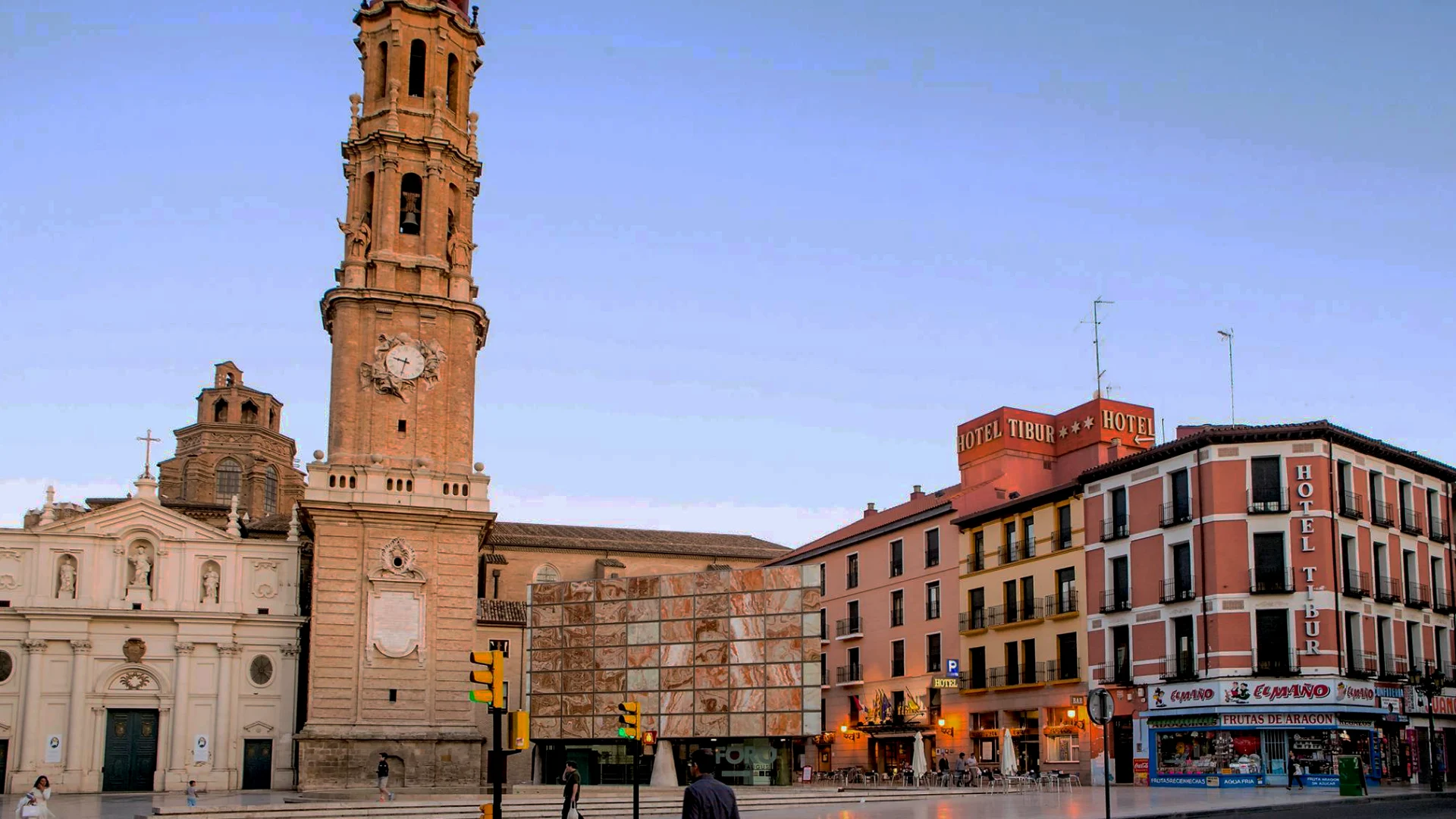 Una plaza con iglesia, torre y un hotel al fondo al atardecer.