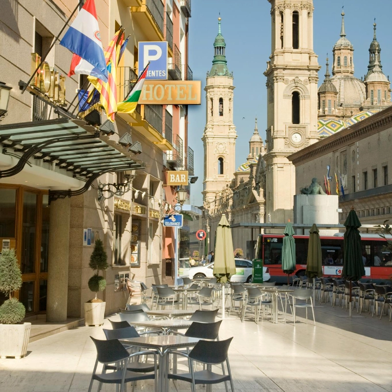 Calle con terrazas al aire libre, hoteles y una catedral al fondo, en un d&iacute;a soleado.
