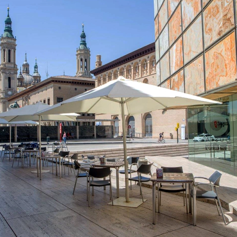 Terraza con sombrillas y mesas en plaza, edificio hist&oacute;rico con torres al fondo, d&iacute;a soleado.