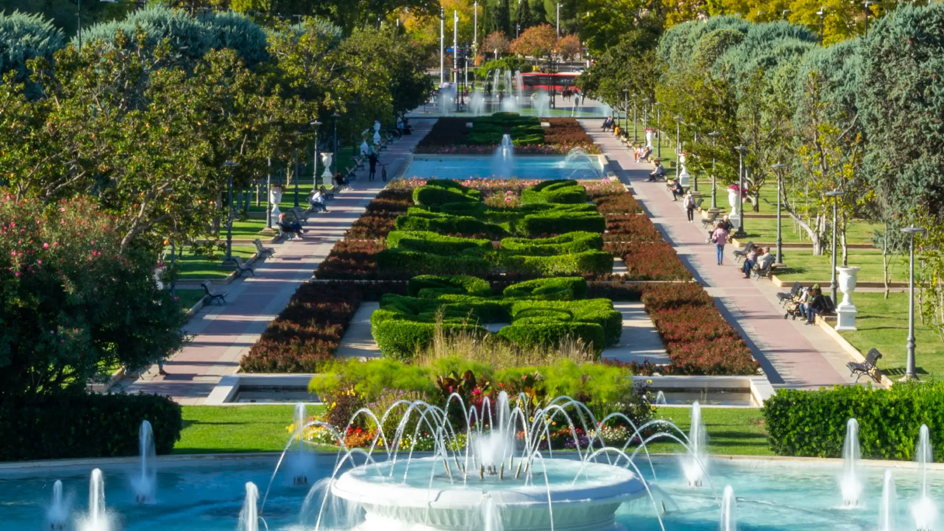 Parque con fuentes, jardines y personas caminando por un sendero rodeado de &aacute;rboles.