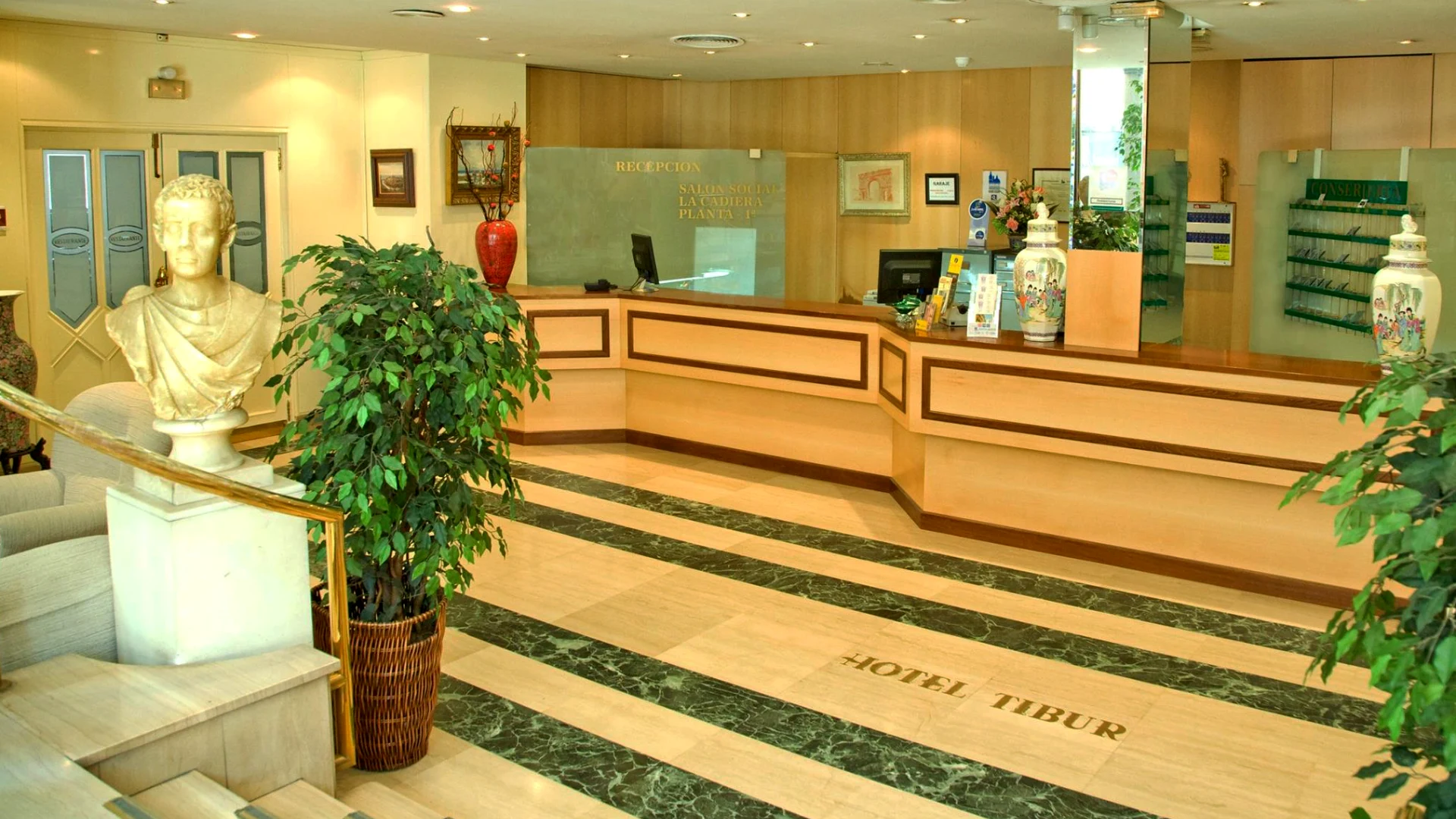 Lobby del Hotel Tibur con mostrador de recepci&oacute;n, plantas y busto decorativo.