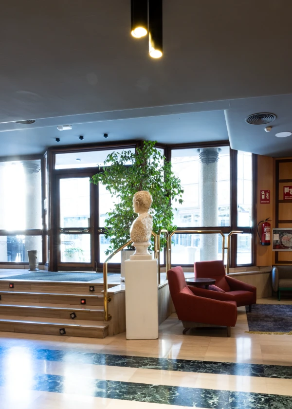 Vest&iacute;bulo elegante con escultura, sillones rojos y planta decorativa.