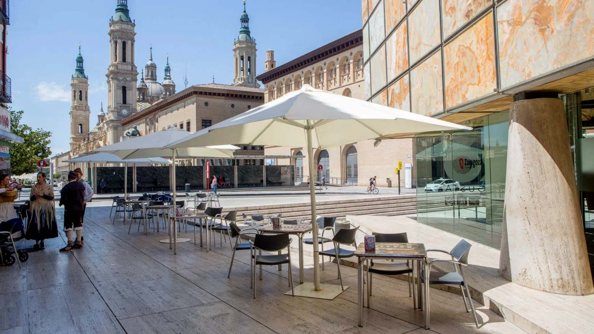 Terraza con mesas y sombrillas frente a edificio hist&oacute;rico con torres al fondo, d&iacute;a soleado.