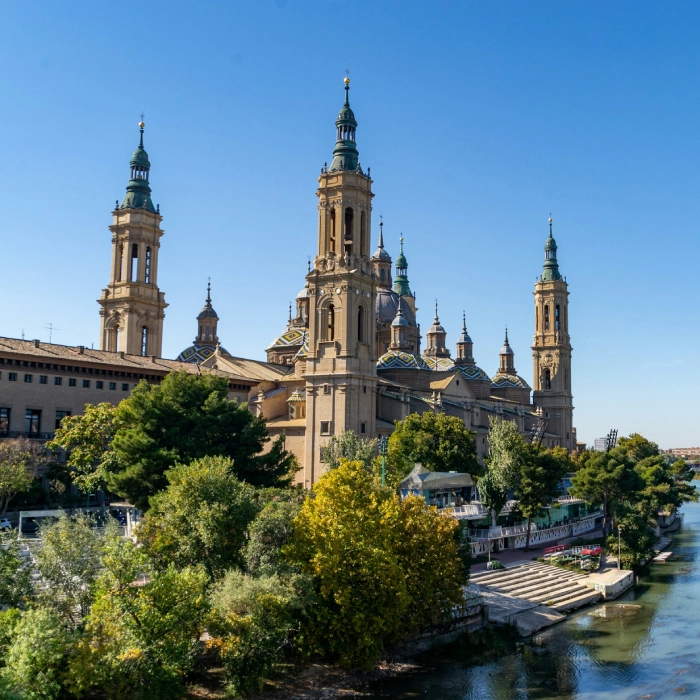 Catedral con torres verdes junto a un r&iacute;o, rodeada de &aacute;rboles y bajo un cielo azul despejado.