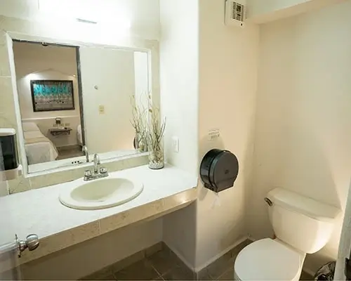 Baño con lavabo blanco, espejo grande, inodoro y puerta que conecta a una habitación con cama.
