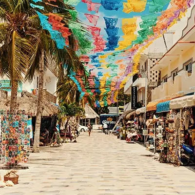 Calle con banderines de colores, palmeras y tiendas, en un ambiente soleado y festivo.