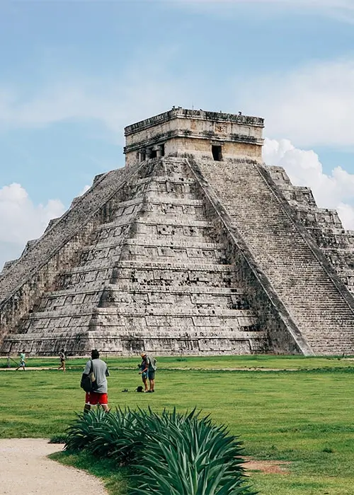Pirámide antigua en un entorno verde, con turistas caminando cerca y cielo azul.