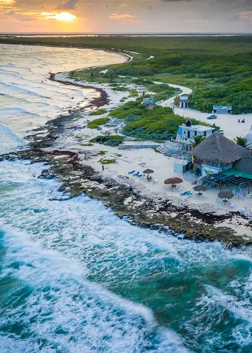 Vista aérea de una playa con olas, vegetación y una cabaña al atardecer.