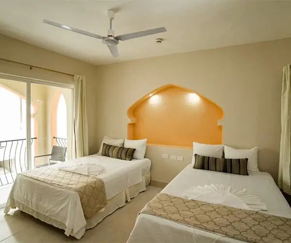 Habitación con dos camas, ventilador de techo y balcón. Decoración en tonos cálidos.