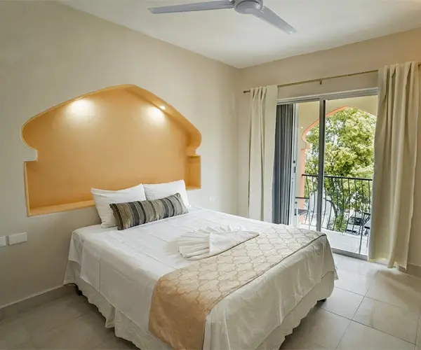 Dormitorio con cama doble, ventanas grandes y decoración simple, vista hacia el exterior.