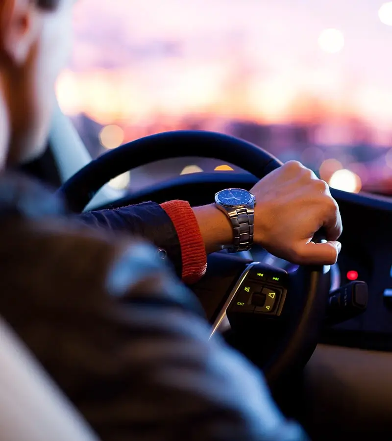 Persona manejando un auto al atardecer, con reloj metálico en la muñeca.