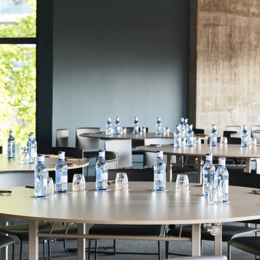 Mesas redondas con botellas de agua y vasos en una sala de conferencias con sillas vacías.