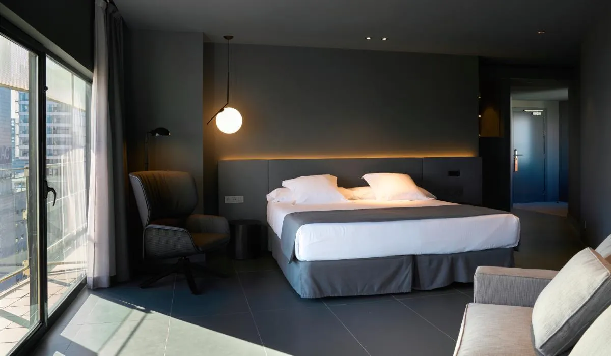 Habitación moderna con cama doble, sillón, lámpara colgante y ventana grande.