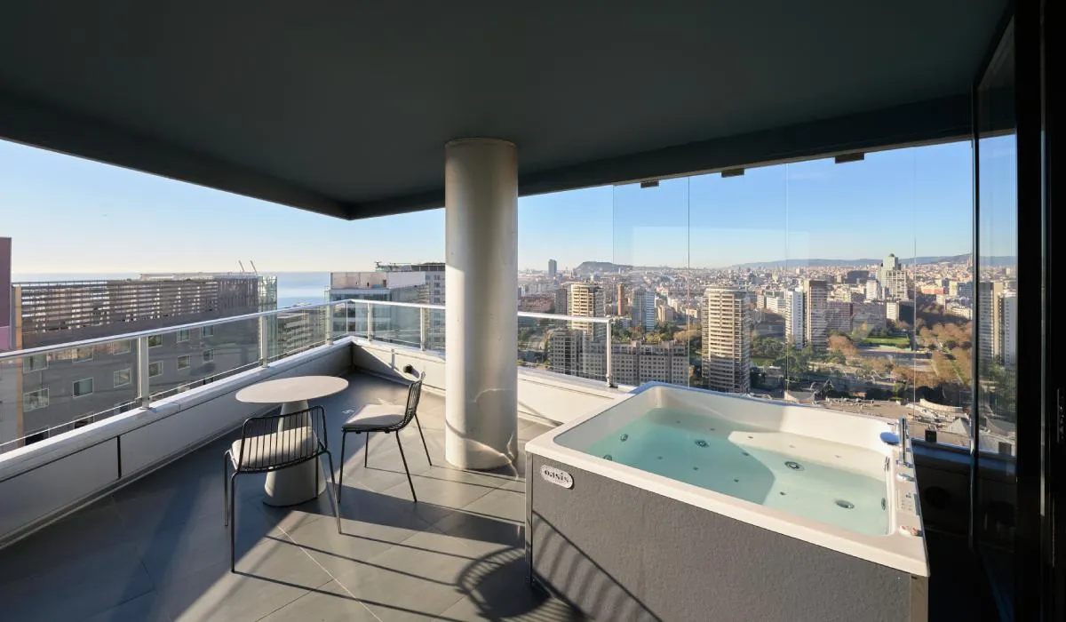 Balcón alto con jacuzzi y mesa, vista panorámica de la ciudad y el mar al fondo.