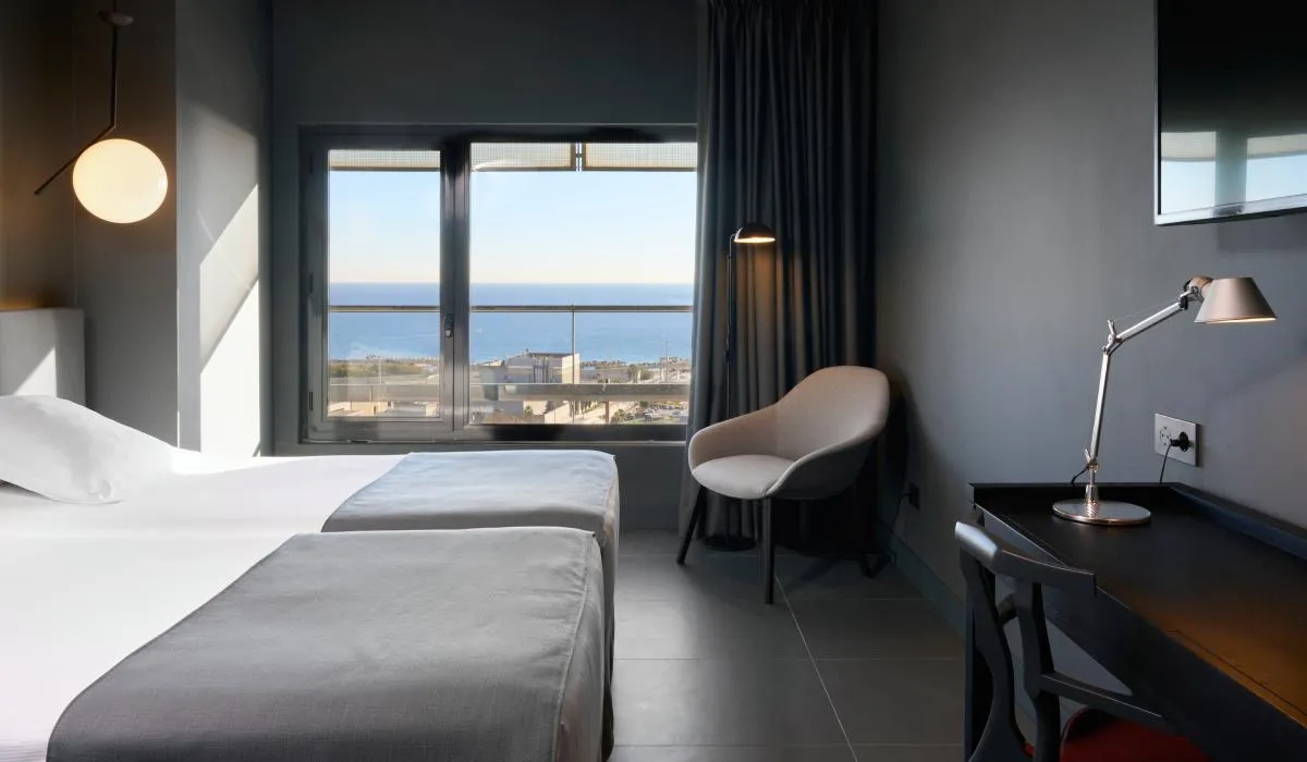 Habitación moderna con dos camas, escritorio, silla y vista al mar por la ventana.