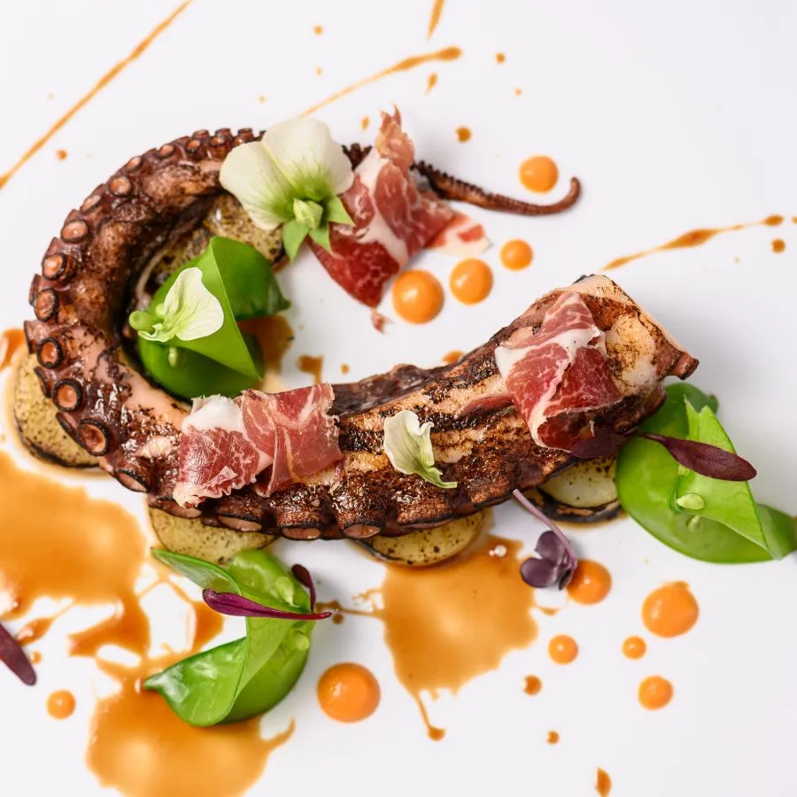 Pulpo a la parrilla con jamón, flores y hojas verdes sobre salsa naranja. Decoración sofisticada en plato blanco.