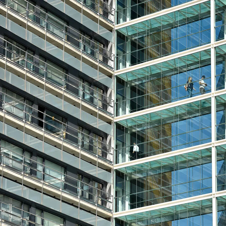 Edificio moderno con fachada de vidrio y balcones; personas caminan en un pasillo elevado de cristal.